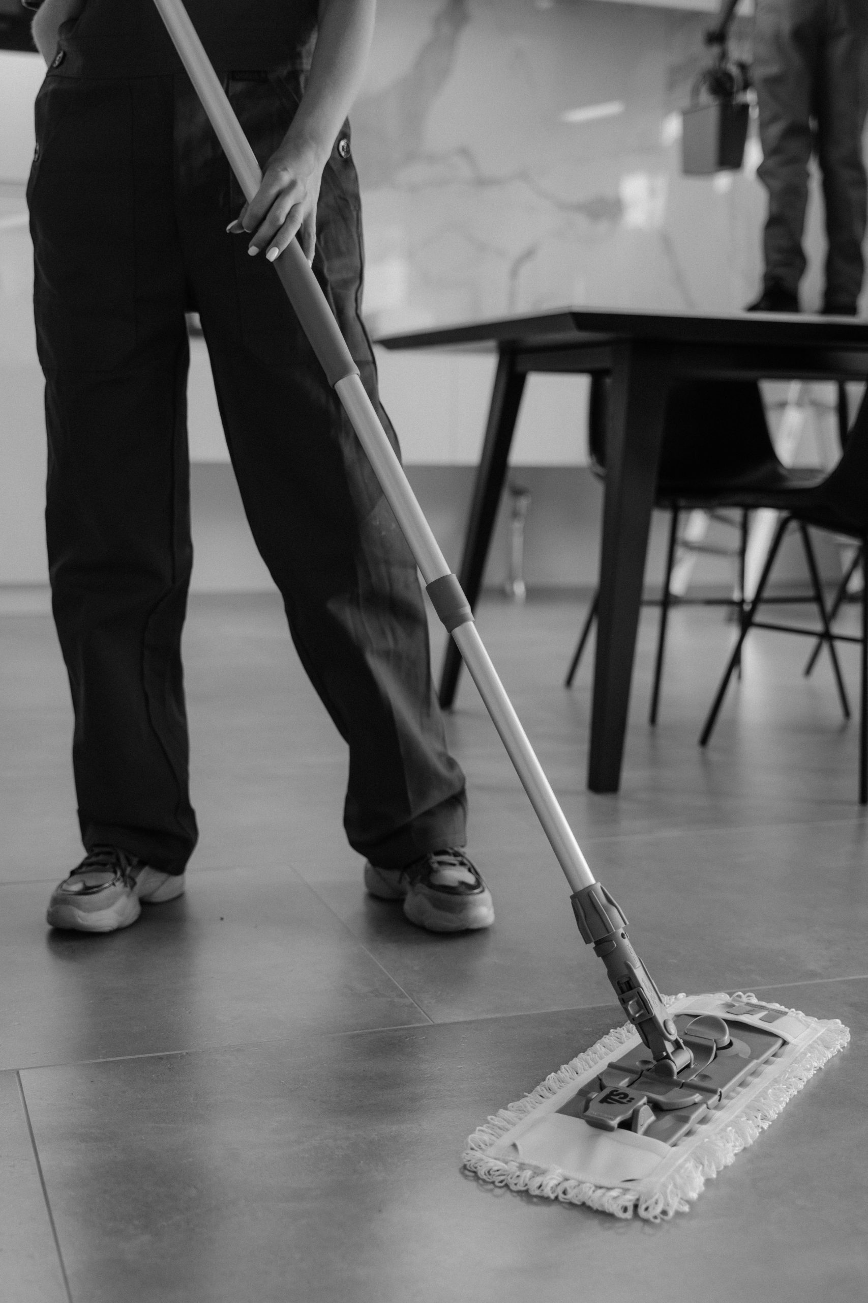 Monochrome image of a person mopping a tiled floor in an indoor setting.